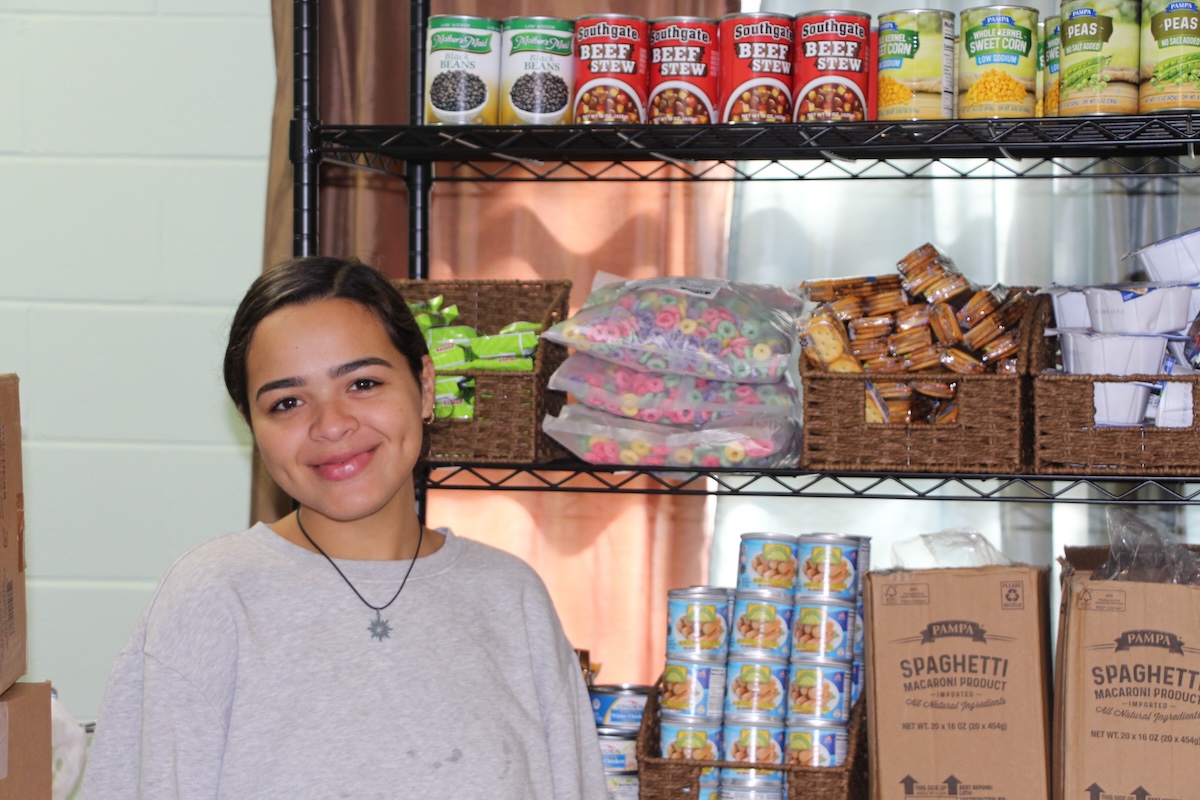 Community Ambassador Samantha Ortega poses in front of a shelf of dry goods at the food pantry she helps run.